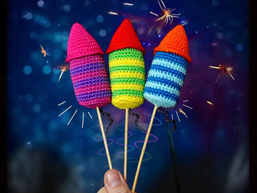 Drei bunte gehäkelte Raketen auf Holzstäben mit gestreiften Mustern und konischen Spitzen, vor funkelndem dunklem Hintergrund, gehalten von einer Hand.