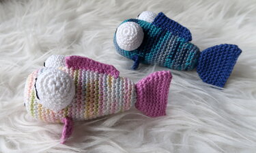 Zwei handgehäkelte Amigurumi-Fische, einer in Pastellfarben mit pinken Flossen, der andere gestreift in Blau- und Türkistönen, auf weißem, flauschigem Hintergrund.