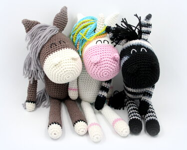 Drei gehäkelte Amigurumi-Stofftiere: brauner Esel, weißes Einhorn mit regenbogenfarbener Mähne und schwarz-weiß gestreiftes Zebra auf hellem Hintergrund.