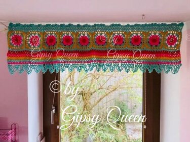 Bunte gehäkelte Fensterblende mit floralen Granny Squares in Pink, Gelb und Türkis, am oberen Fensterrand angebracht.