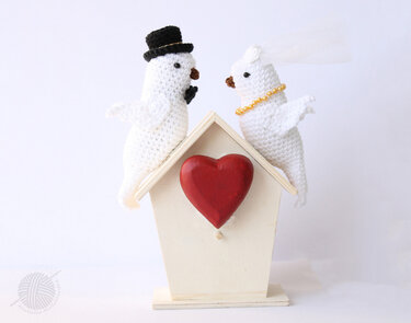 Two white crocheted birds (bride and groom) perched on a wooden birdhouse with a red heart.