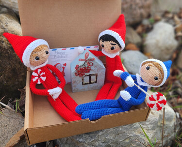 Three crocheted elf dolls in red and blue sitting in a cardboard box with a holiday card, small house ornament and peppermint lollipops