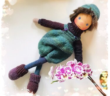 Gehäkelte Amigurumi-Puppe im grünen Latzkleid mit Mütze und violetten Schuhen neben einer Orchideenblüte