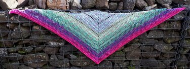 Gehäkeltes Dreieckstuch in Pink, Lila, Türkis und Grün mit Lochmuster, auf einem Gabionenkorb drapiert.