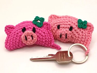 Zwei gehäkelte rosa Schweinchen‑Schlüsselanhänger mit grünen Häkelkleeblättern neben einem Schlüssel und Schlüsselring