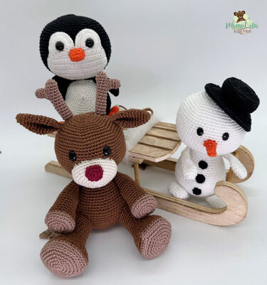 Gehäkelter Pinguin hinter einem gehäkelten Rentier und neben einem gehäkelten Schneemann mit schwarzem Hut, davor ein kleiner Holzschlitten
