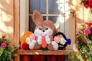Crochet bunny holding a carrot between two floral eggs on a wooden doorstep