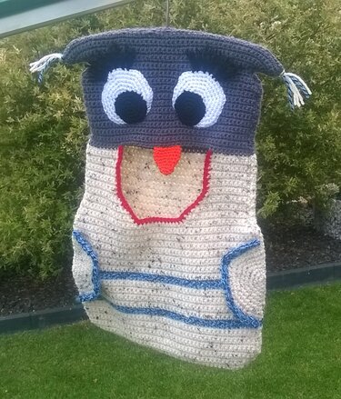 Gehäkeltes Eulen-Utensilo in Grau-Beige mit großen Augen und orangefarbenem Schnabel, hängend im Garten