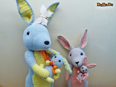 Zwei gehäkelte Amigurumi-Hasen in Pastellfarben, jeder hält ein kleines Babyhäschen; blau-gelber Hase mit Schleife und rosa-grauer Hase