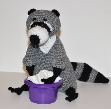 Gehäkelter Amigurumi-Waschbär grau-schwarz mit gestreiftem Schwanz und lila Schüssel