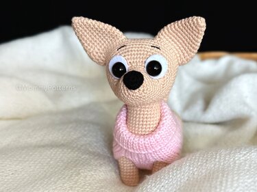 Tan crocheted chihuahua amigurumi with large ears wearing a pink sweater seated on a cream blanket