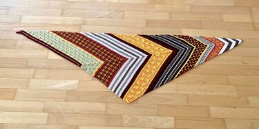 Dreieckiges Stricktuch mit komplexen Mustern in Braun, Rot, Gelb und Grau auf hellem Holzuntergrund.