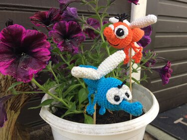 Zwei gehäkelte Insekten-Amigurumi (orange und blau) als Pflanzenstecker in einem weißen Blumentopf mit violetten Petunien.