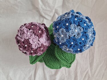 Zwei gehäkelte Hortensienblüten in Blau und Lila mit Perlen und grünen Blättern.