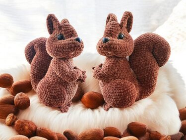 Zwei braune gehäkelte Eichhörnchen-Amigurumi auf weißem Fell, Nüsse liegen rundherum