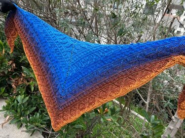 Crochet triangle shawl with blue-to-orange gradient and textured stitch pattern, displayed outdoors.