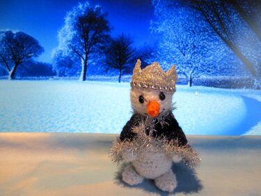Gehäkelter Amigurumi‑Schneemann mit silberner Krone und Lametta‑Schal vor winterlicher Kulisse