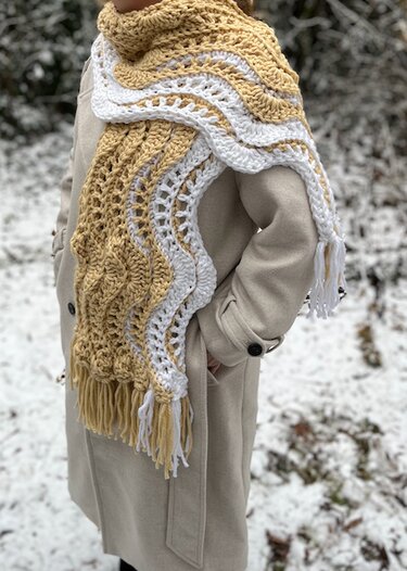 Two-tone beige and white crocheted scarf with wavy openwork pattern and fringe worn over a coat in snow.