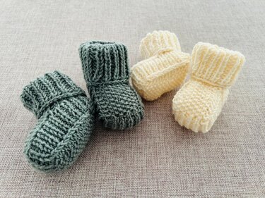 Zwei grüne und zwei cremefarbene gestrickte Babyschuhe liegen auf einem Textiluntergrund