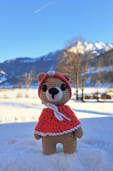 Gehäkelter brauner Amigurumi-Teddybär mit roter Kapuze und weißem Rand vor verschneiten Bergen