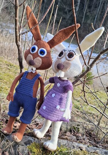 Zwei gehäkelte Amigurumi-Hasen, braun mit blauer Latzhose und weiß mit lila Kleid, vor kahlen Ästen im Freien