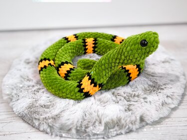 Gehäkelte grüne Amigurumi‑Schlange mit gelb‑schwarzen Streifen auf rundem Fellkissen