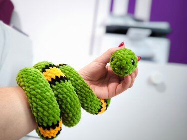 Hand hält eine gehäkelte grüne Schlange mit gelben und schwarzen Streifen, gewickelt ums Handgelenk.