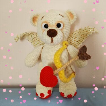 Cream crocheted amigurumi teddy bear with gold-sequined wings holding a yellow bow and a red heart-tipped arrow, red paw pads visible
