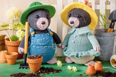 Zwei gestrickte Maulwürfe: links mit blauer Latzhose und grünem Hut, rechts mit hellblauem Kleid und gelbem Hut, dekoriert mit Tontöpfen, Erde und Gartenwerkzeug.