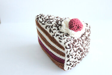 Gehäkeltes Kuchenstück mit weißen und braunen Schichten und rosa Kirsche oben