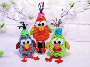 Three crocheted amigurumi birds in gray, orange and green with large white eyes, orange beaks, red crocheted feet and yarn tufts on a pale fabric background.