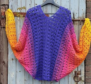 Gehäkelter Damenponcho mit Farbverlauf in Lila, Pink, Orange und Gelb, mit durchbrochenem Muster und V-Ausschnitt, hängend vor einer hellblauen Holztür.