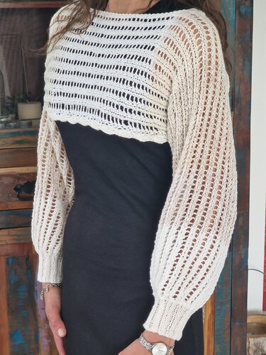 Person trägt cremefarbenen kurzgestrickten Bolero mit Ajourreihen und langen Ballonärmeln über schwarzem Kleid