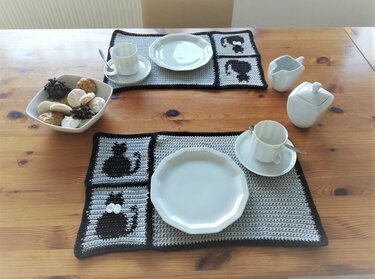 Gehäkeltes graues Tischset mit schwarzem Rand und Katzen-Motiven, Holztisch mit weißem Geschirr