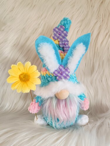 Gehäkelter Pastell-Wichtel mit Hasenohren, flauschigem Bart, gelber Blume und Mütze mit Stoffblüten vor hellem Hintergrund