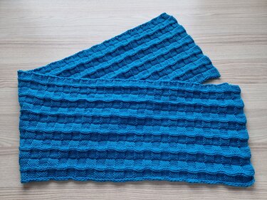 Blauer gestrickter Schal mit reliefartigem Blockmuster auf hellem Holzuntergrund