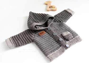 Gehäkelte graue Kinderjacke mit Kapuze, Rippenbündchen und Knopfleiste auf weißem Hintergrund, daneben ein Holzspielzeug.