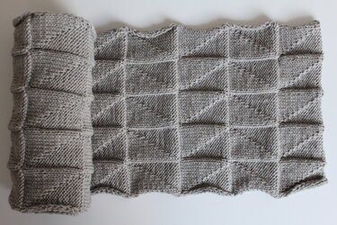 Grauer gestrickter Schal mit quadratischem Reliefmuster, teilweise aufgerollt auf hellem Hintergrund