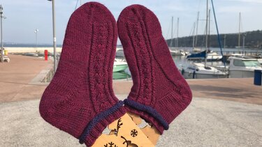 Paar bordeauxrote, handgestrickte Socken mit Zopfmuster auf Holzspanner vor Bootshafen