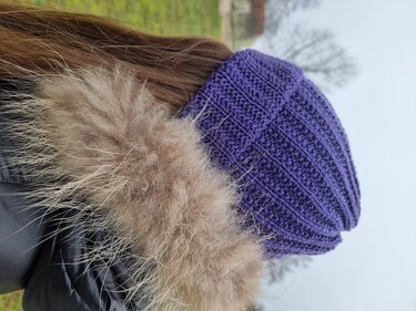 Dunkelviolette Strickmütze mit Rippenmuster, seitliche Ansicht neben Mantel mit hellem Fellbesatz