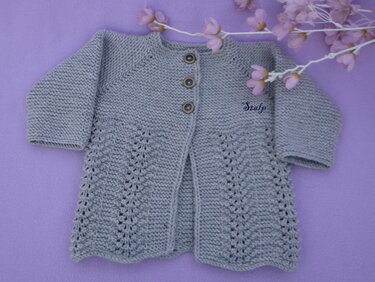 Graue, handgestrickte Babyjacke mit Lochmuster und drei Holzknöpfen, liegend auf lila Stoff mit zarten Blüten.