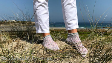Füße in weißen Hosen mit kurzen gestrickten Pastellsocken auf sandigen Dünen vor dem Meer