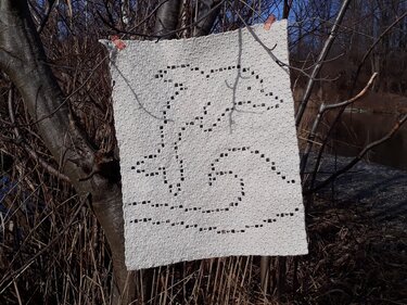 White filet crochet square showing two jumping dolphins over a wave, clipped to tree branches