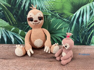 Zwei gehäkelte Amigurumi-Faultiere in Braun und Beige, ein großes und ein kleines, auf Holz vor grüner Blätterkulisse.