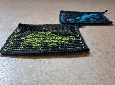 Zwei gestrickte Topflappen auf hellem Untergrund: einer mit grünem Baum- bzw. Drachenmotiv auf dunklem Grund, der andere mit blauem Krabben- bzw. Hummermotiv.