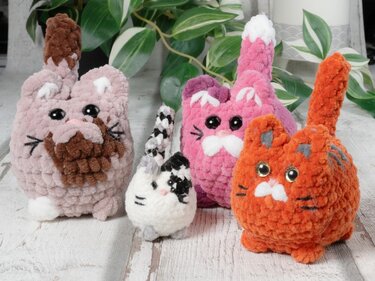Vier flauschige gehäkelte Amigurumi-Katzen in Rosé, Pink, Orange und Weiß-Schwarz auf hellem Holztisch.