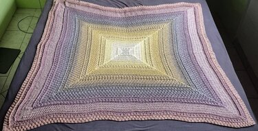 Quadratische Häkeldecke mit sanftem Verlauf von Hellgelb zu Blau-, Lila- und Rosatönen auf dunkelgrauem Untergrund.