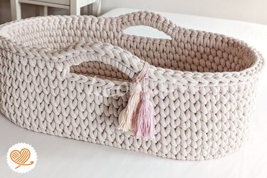 Ovaler gehäkelter Babykorb in hellem Beige mit eingearbeiteten Henkeln und zwei pastellfarbenen Quasten