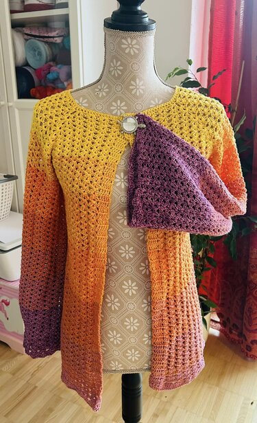 Lange gehäkelte Damenjacke in Gelb, Orange und Lila auf einer Schneiderpuppe mit angeheftetem lila Accessoire.
