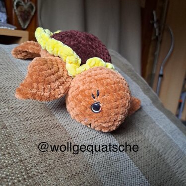 Gehäkelte plüschige Amigurumi‑Schildkröte in Orange, Braun und Gelb, liegend auf Stoff